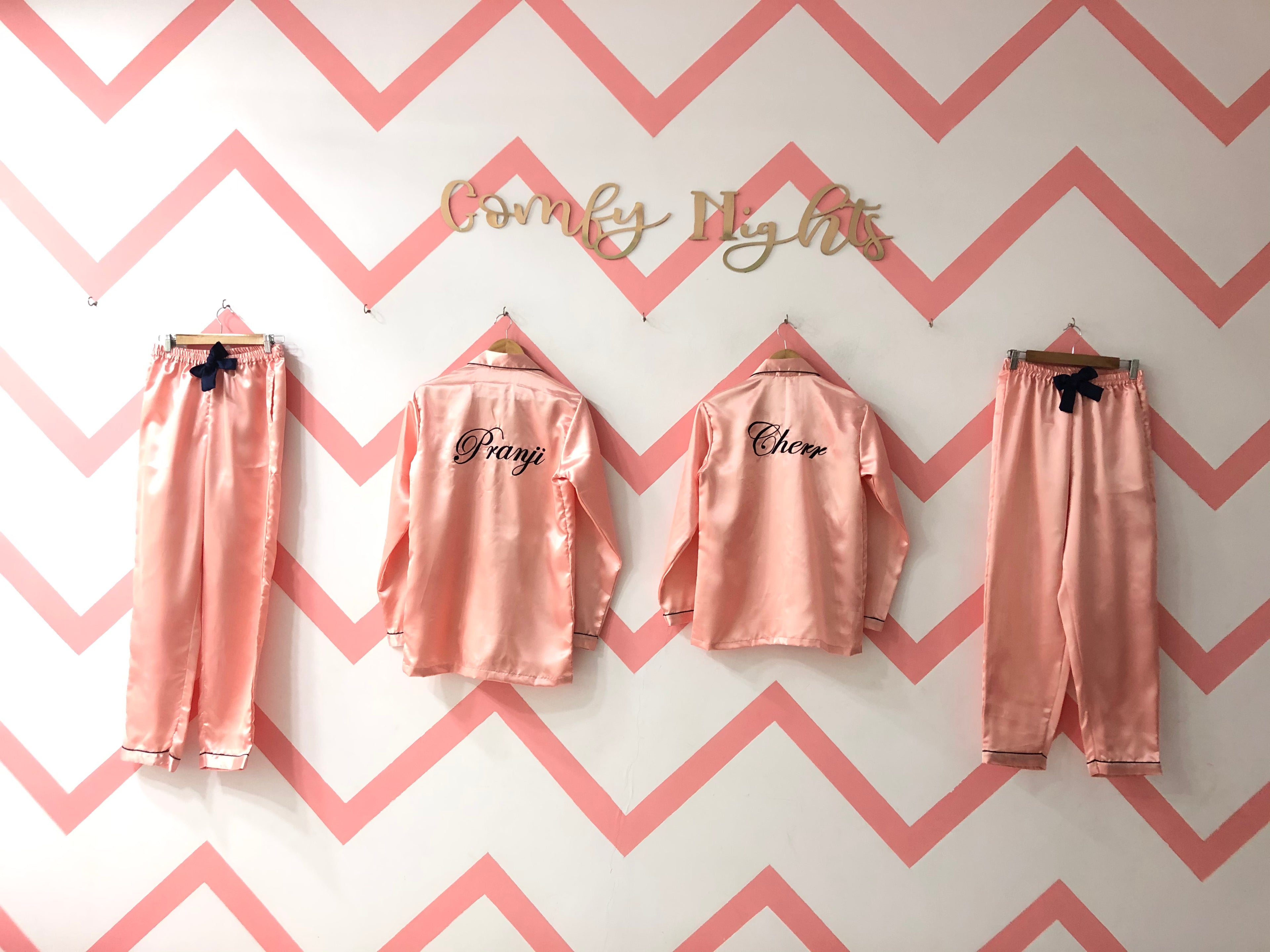 A couple of pink satin pajama sets hanging on hangers against a chevron patterned wall, with the names 'Danny' and 'Claire' printed on the back of the shirts.